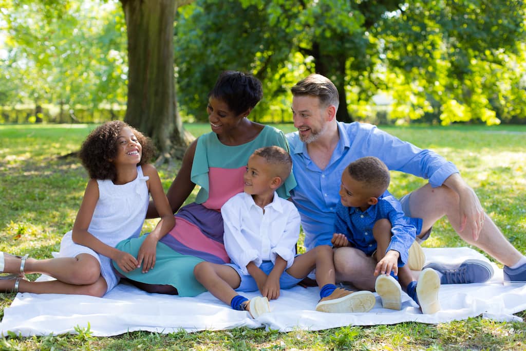 A family formed of two adults and three children are sitting outside in the park on a picnic blanket