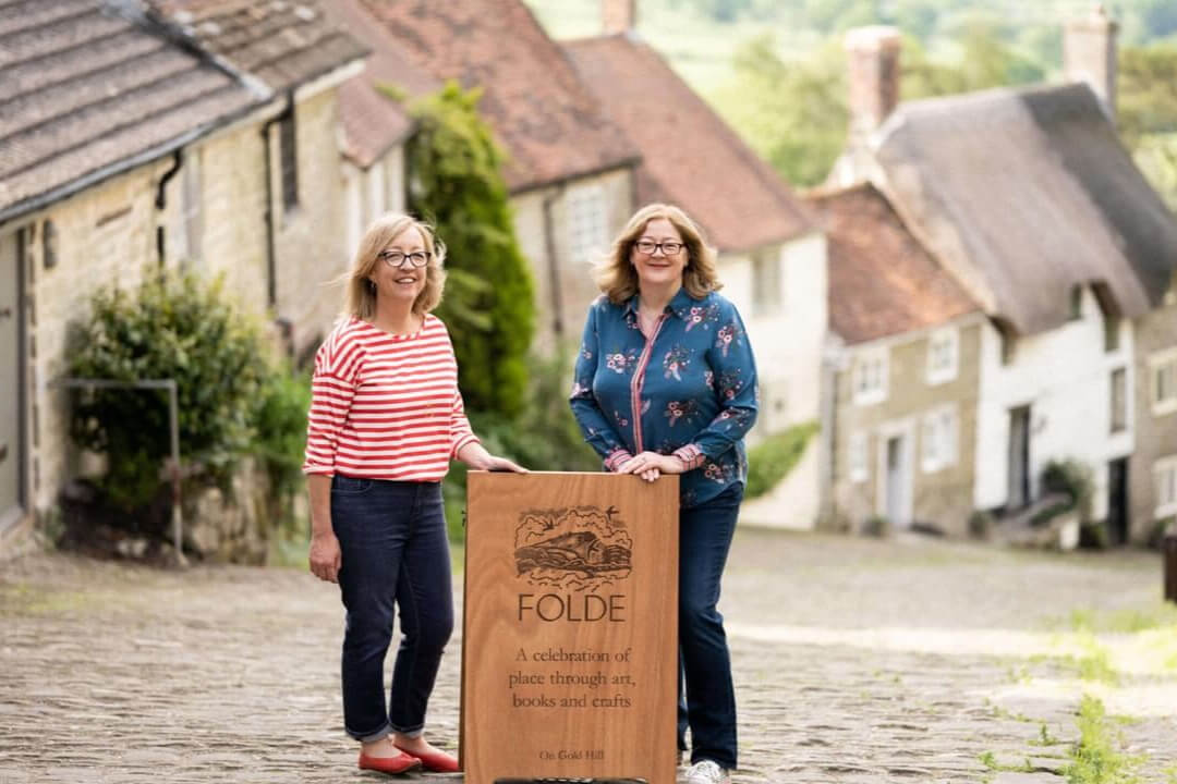 Two women at the top of Gold Hill in Shaftsbury by a shop sign that reads FOLDE