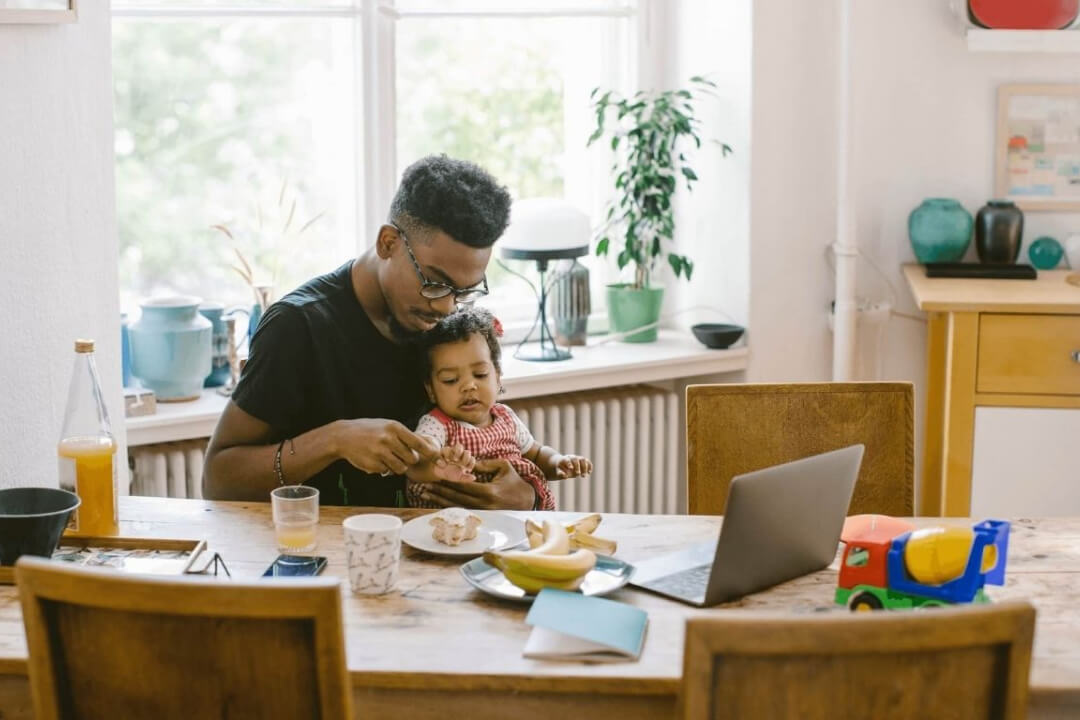 Father sits at a kitchen table with his daughter and an open laptop