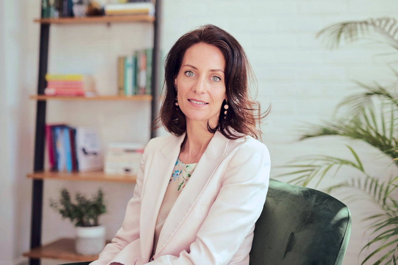 Woman sitting in a green chair indoors with a bookshelf and plant in the background