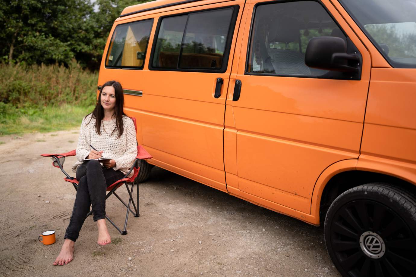 Charlotte sits in a camping chair with a pan and notepad near the orange campervan.