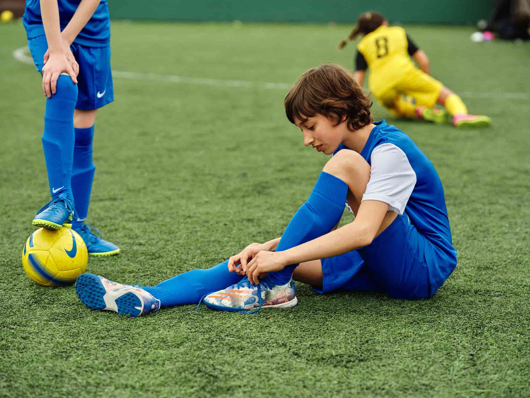 A girl sitting on the grass tying the laces of a football shoe.