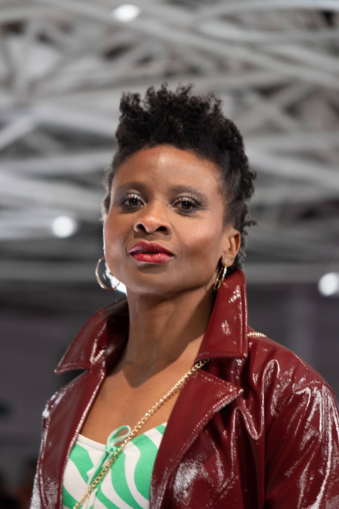 Woman in red leather jacket looks to camera, she is on a catwalk and wearing gold hoops