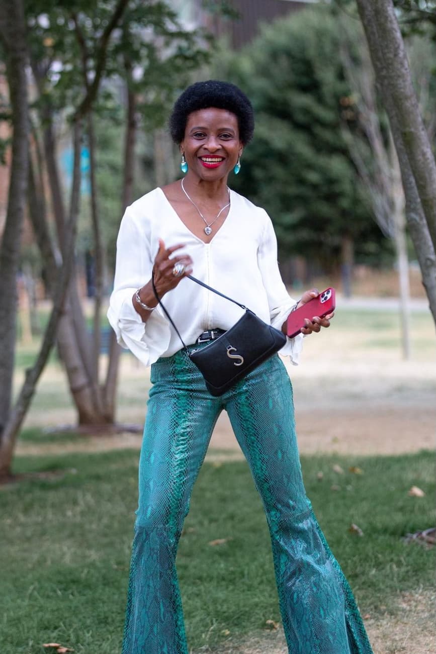 Woman in white shirt and flares looks to camera smiling, she is outside in a park
